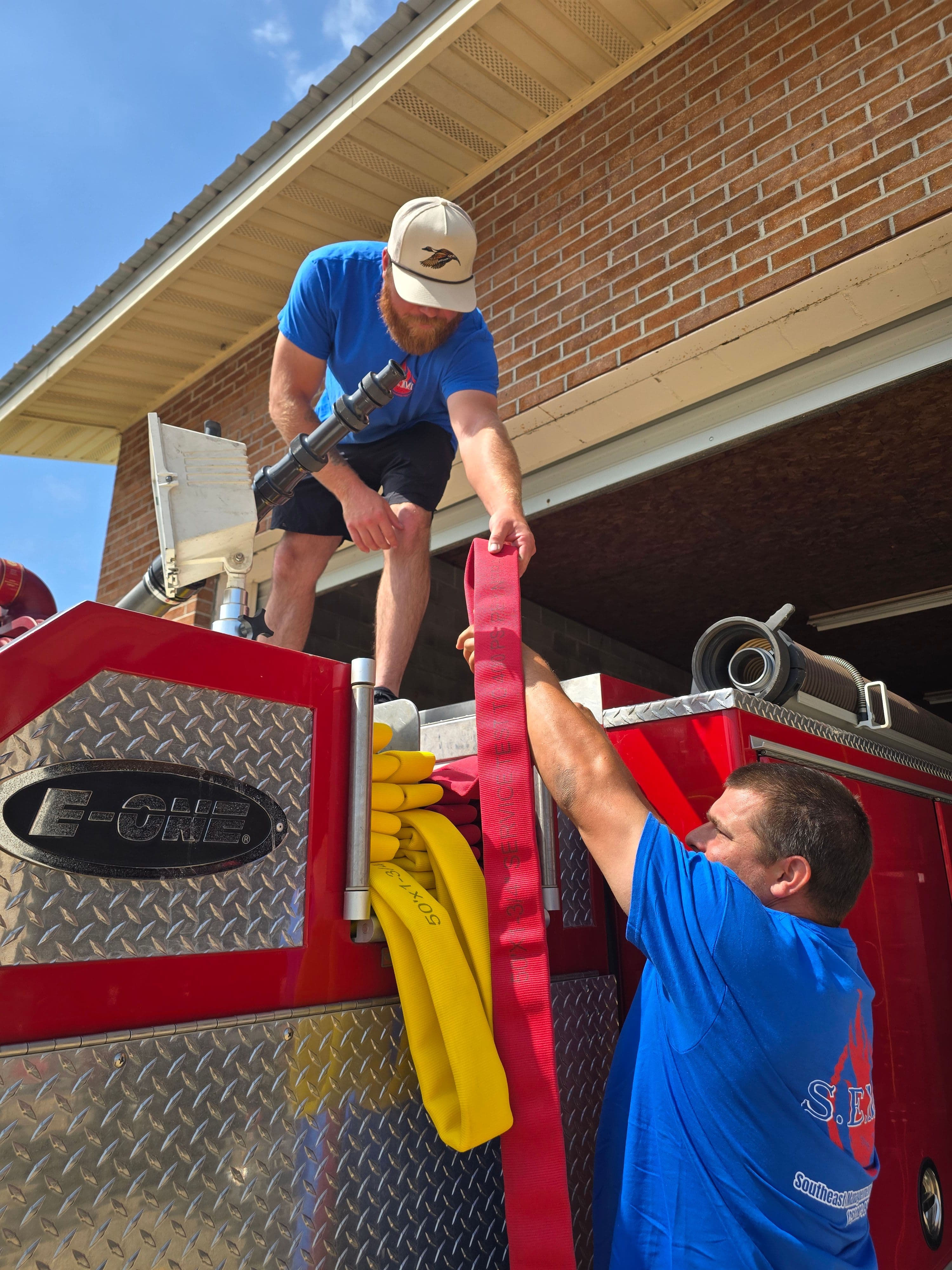 Two SEMS technicians reloading supply hose onto a red E-One fire engine outside the station