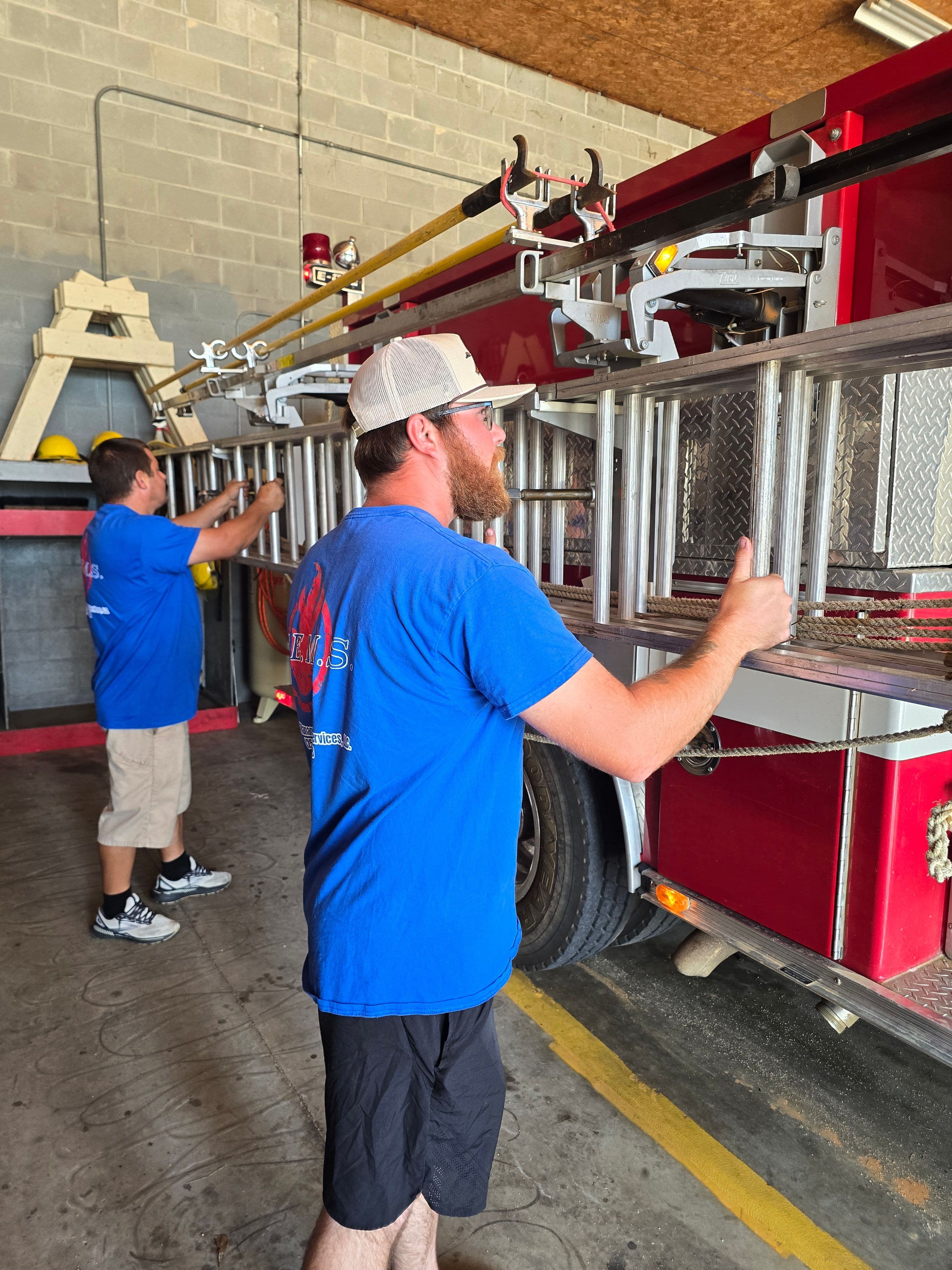 SEMS technicians handling ground ladders mounted on the side of a red fire apparatus in the bay